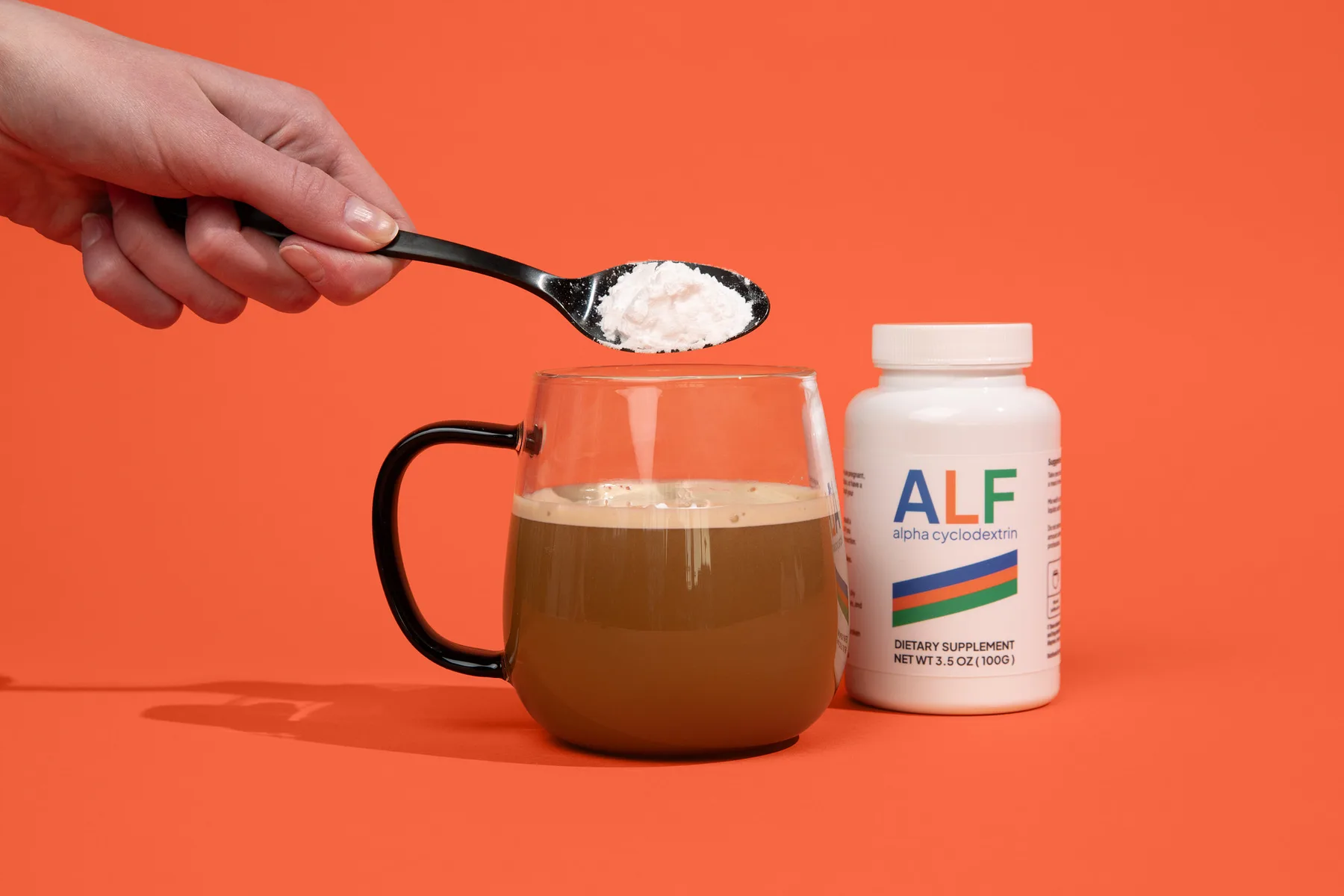 A scoop of ALF Alpha Cyclodextrin powder being added to a mug of coffee, with the bottle beside it